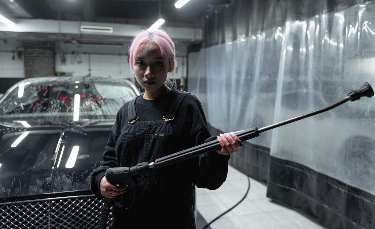 A woman with pink hair uses a pressure washer in a car wash setting. Indoor, nighttime shot.