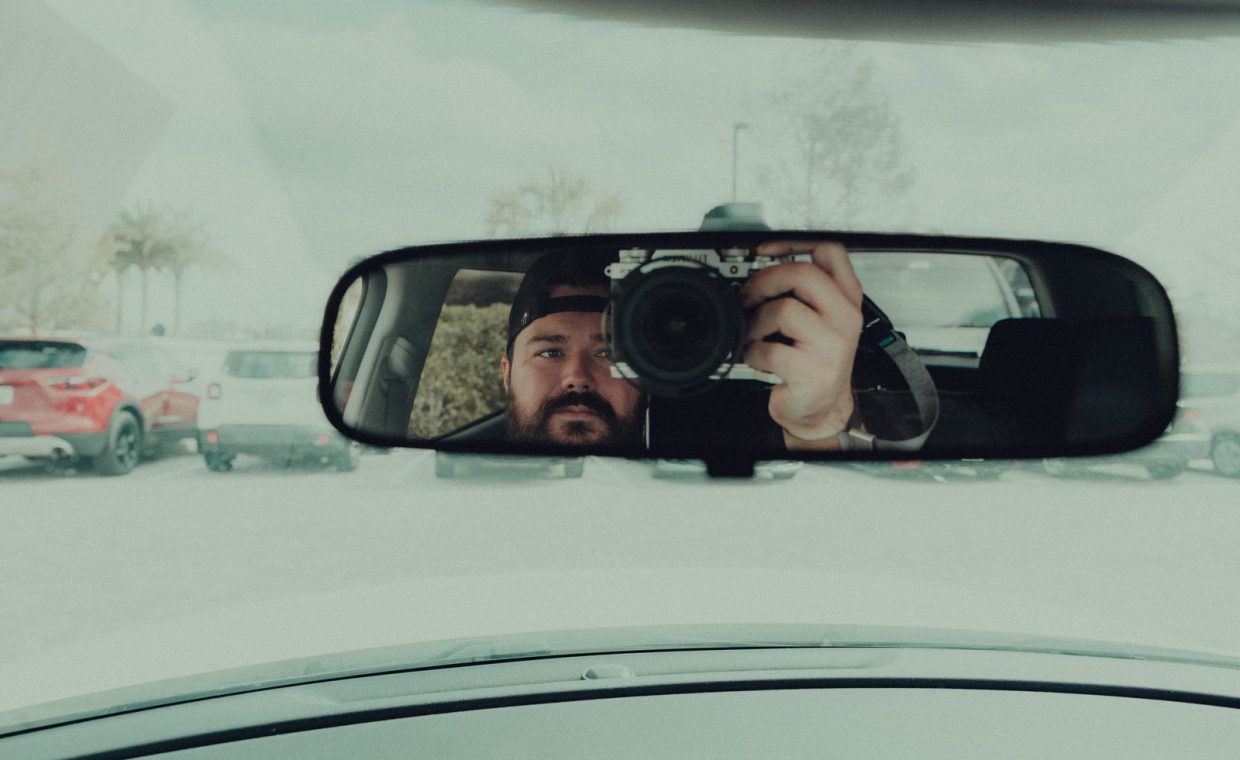 A man captures his reflection in a car's rearview mirror using a camera in a parking lot.