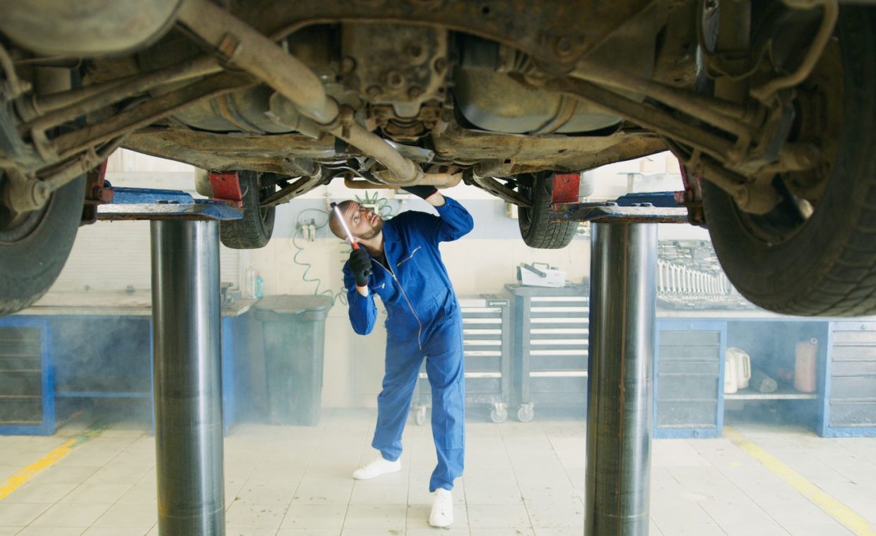 A mechanic in blue uniform inspects a car lifted in a garage workshop.